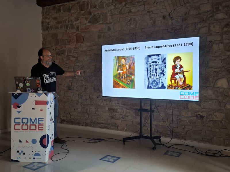 Un relatore accanto al leggio di Come To Code, con sullo schermo una slide su Henri Maillardet e Pierre Jaquet-Droz