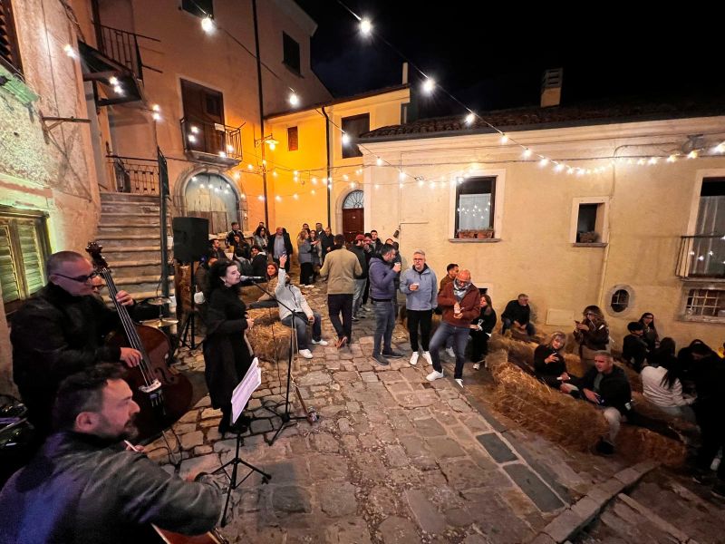 Una serata in una piccola piazza in pietra illuminata da lucine, con musicisti in primo piano e persone in piedi o sedute su balle di fieno