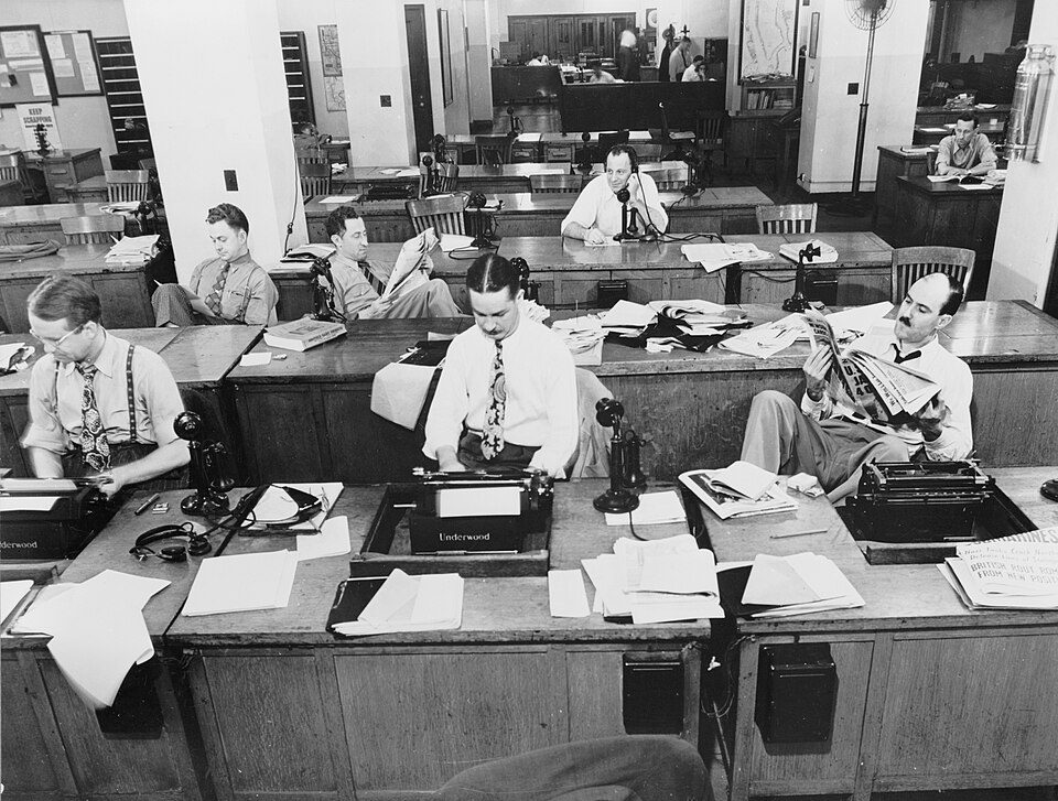 Black-and-white photo of The New York Times newsroom in 1942: men in shirts and ties at rows of wooden desks with typewriters and candlestick telephones, reading newspapers and making calls