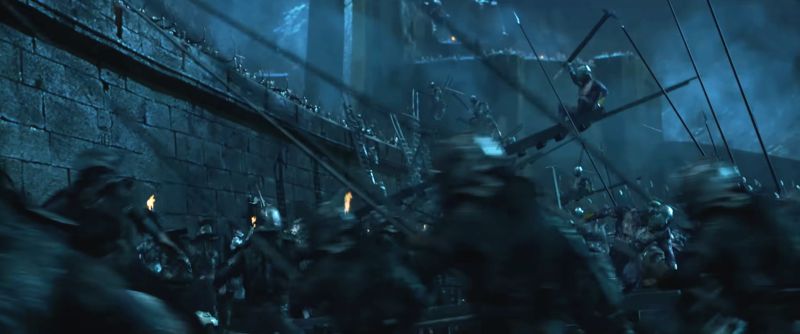 A dark, blurred scene of armoured soldiers storming a fortress wall at night, lit by torches