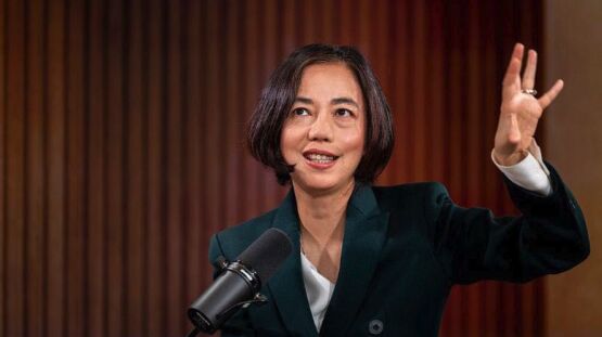A woman in a dark blazer speaking into a studio microphone with one hand raised